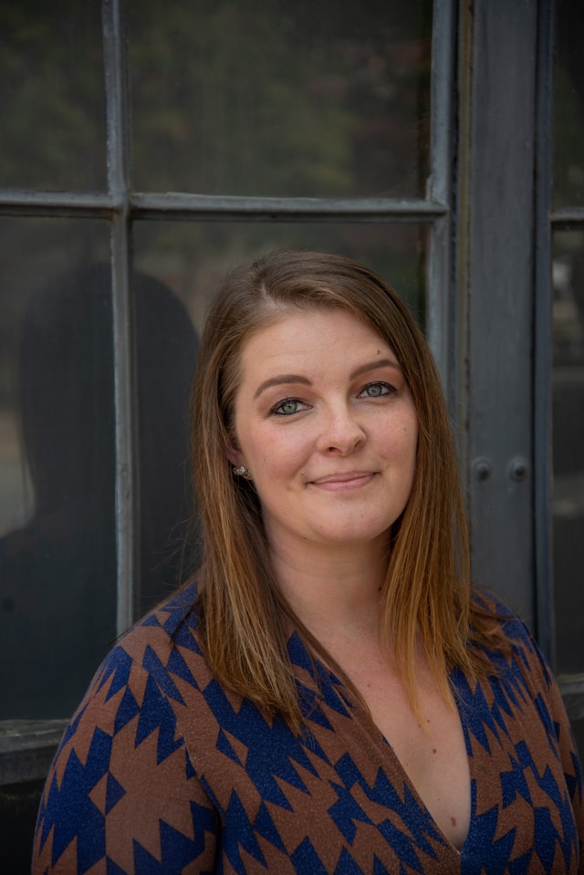 Smiling woman in a patterned shirt, standing by a window.