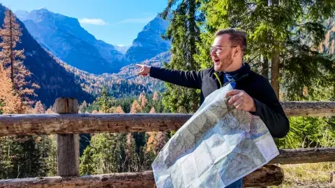 Man with a map points excitedly at mountains in a sunny forest setting.