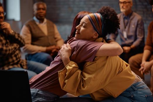 Two women warmly hugging in a supportive group setting.