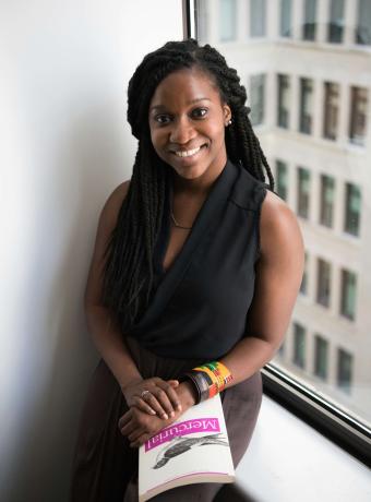 Smiling woman with long hair holds a book near a window.