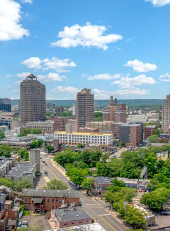 City skyline with tall buildings, trees, and a blue sky with clouds.