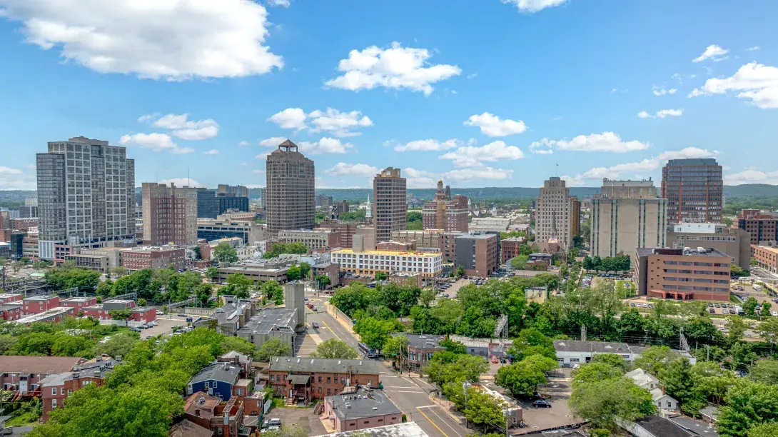 City skyline with tall buildings, trees, and a blue sky with clouds.